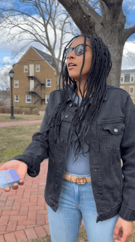 A woman wearing sunglasses and a black jacket stands outside holding a phone, with buildings, trees, and a lamppost in the background.