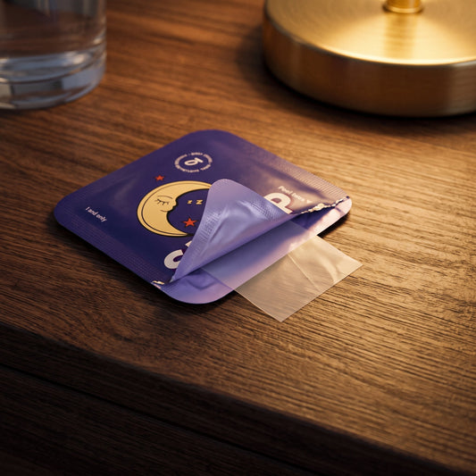 Purple Sleep Melatonin oral strip packet peeled open on a wooden nightstand beside a lamp and glass of water.