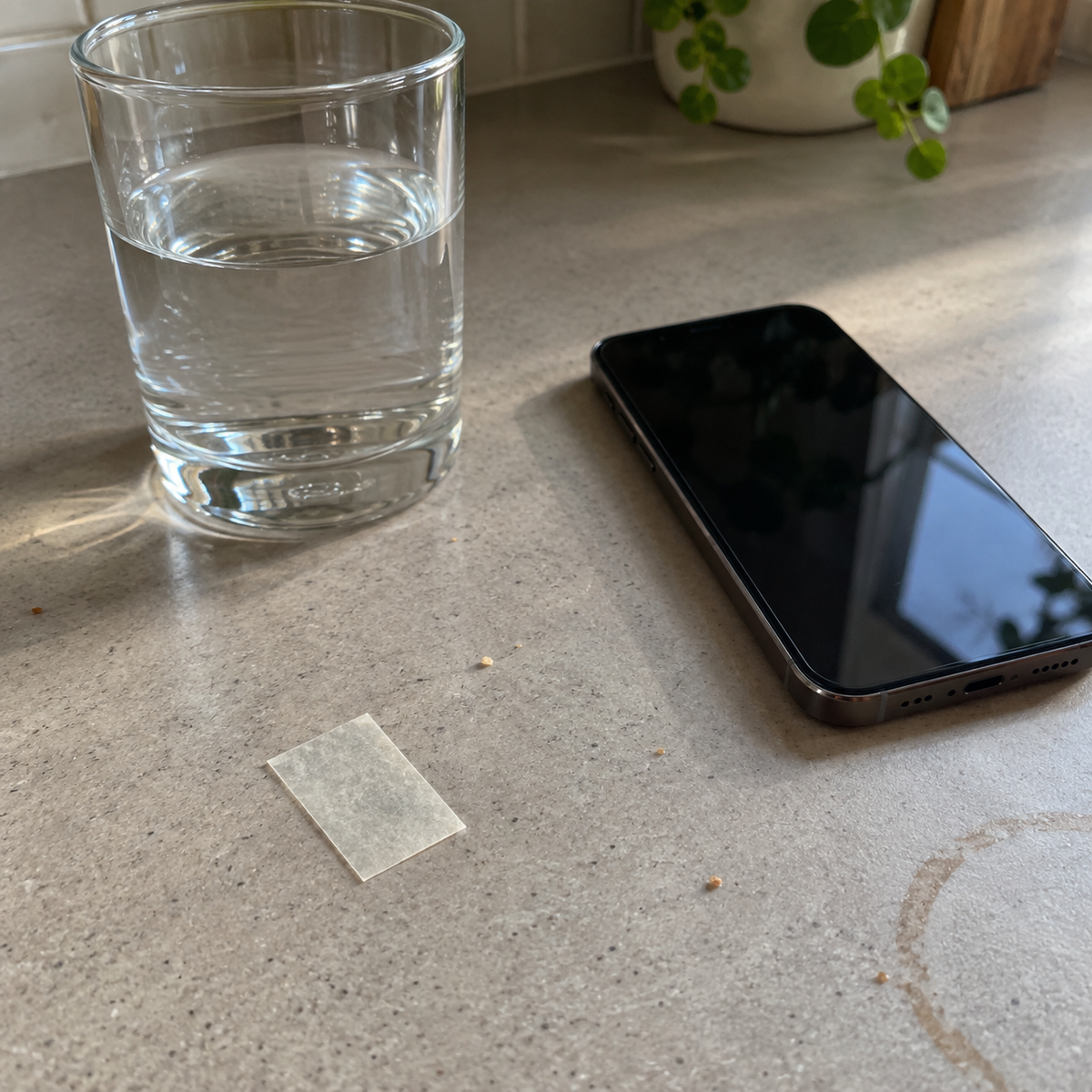 A single thin, translucent dissolvable vitamin strip resting on a kitchen counter next to a phone and a glass of water, no packaging in view.