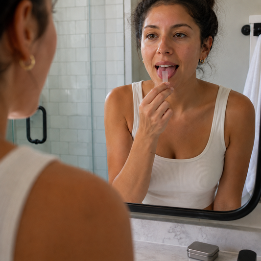 A woman standing at a bathroom mirror in soft morning light, placing a Holy Strips multivitamin oral strip on her tongue as part of her morning routine.