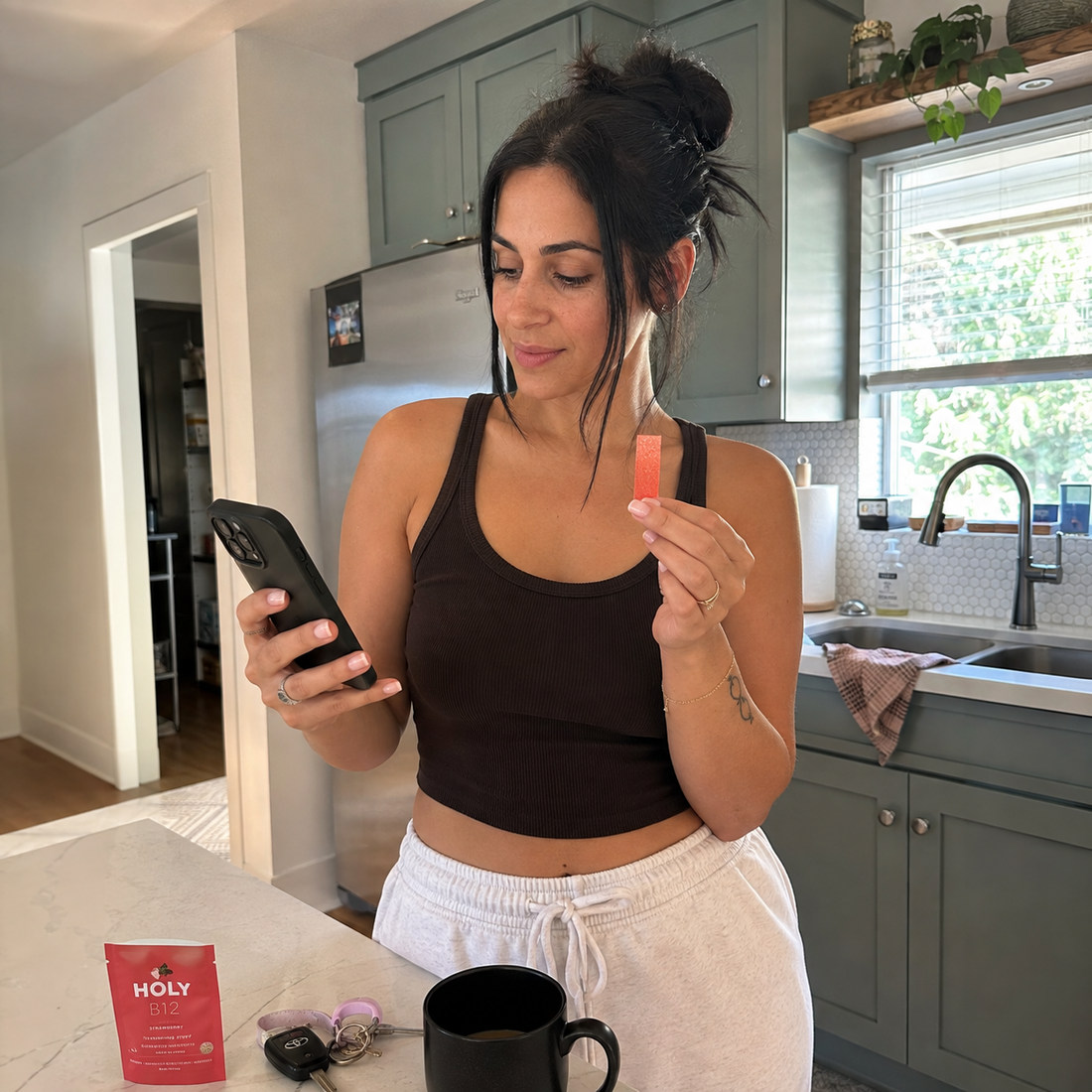 A woman checking her phone while holding a Holy Strips B12 strip at her kitchen counter.