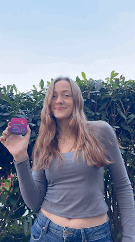 A woman with long hair stands outdoors in front of bushes, holding up a small purple container and smiling.