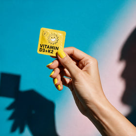A hand with yellow nail polish holds a yellow HolyStrips® D3 + k2 packet, offering an immune boost against a blue background with shadows.