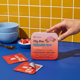 Hand holding a tin labeled “HolyStrips® Vitamin B12” by Holy Strips, a vegan strawberry-flavored energy supplement, beside open packets and strawberries on yellow tiles.