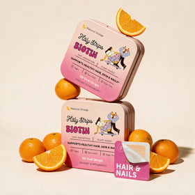 Two pink tins of Holy Strips HolyStrips® Biotin fast-dissolving oral vitamin strips are stacked among whole and halved oranges. A sample strip and “Hair & Nails” card are displayed in the foreground, while the tins feature illustrations of women exercising to emphasize effective support for hair, skin, and nails.
