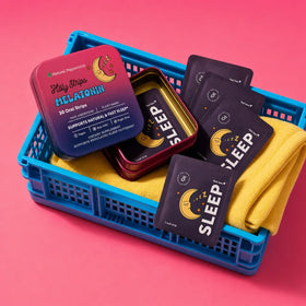 A blue basket lined with a yellow cloth contains a tin of HolyStrips® Melatonin Strips by Holy Strips, labeled “Sleep,” along with six individual melatonin strip packets, all displayed on a pink background.